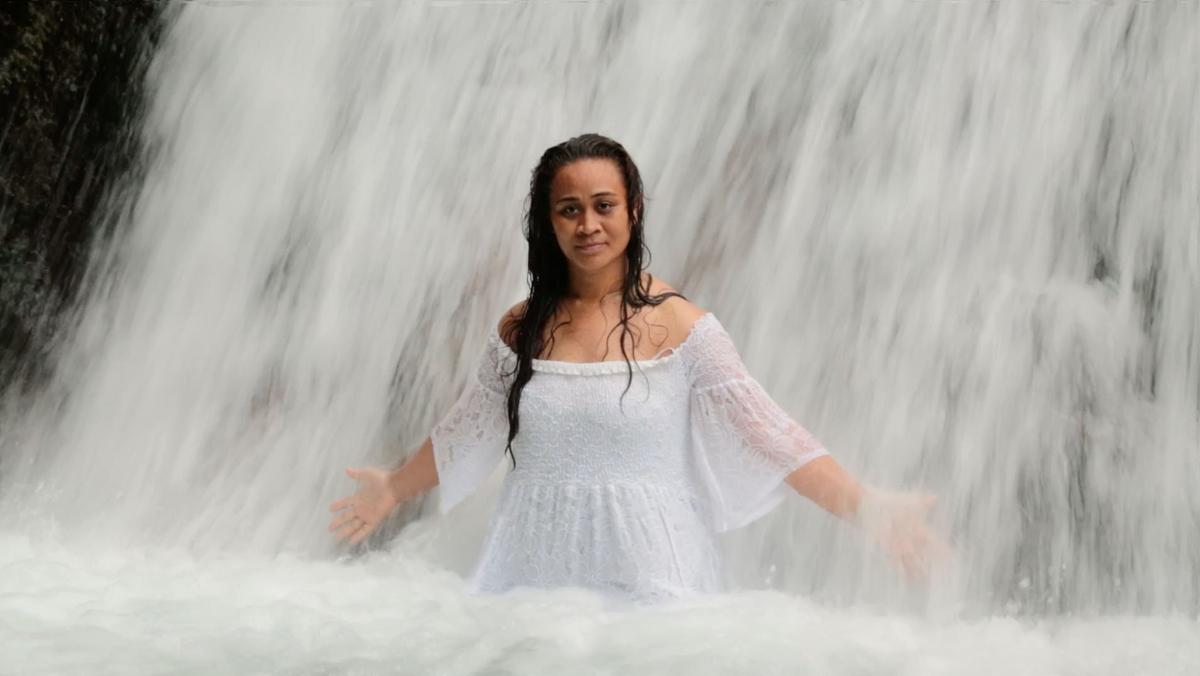 A woman in a white dress stands with her arms open under a waterfall. The white water is cascading over her, she looks serious but calm